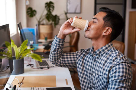 A young Asian businessman drinks coffee at his desk in an empty office