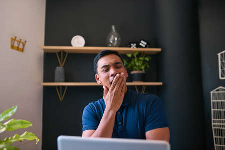 A young Asian man yawns at his desk in the office with his hand over mouth