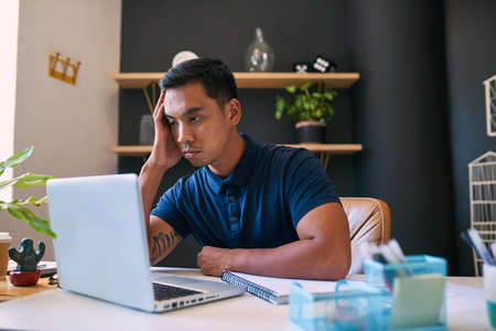 A young Asian man looks bored in the office looking at his computer screen