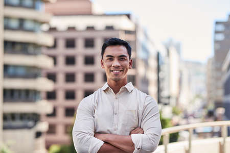 A young attractive Asian man stands in front of city buildings with arms crossedの写真素材