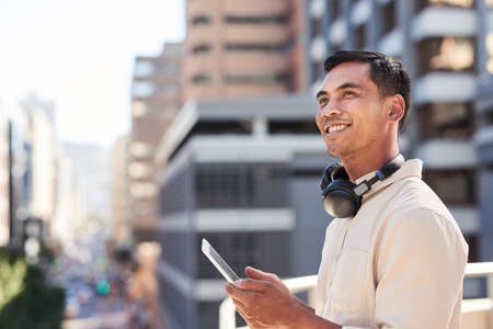 A trendy Asian businessman stands on city balcony with tablet and headphonesの写真素材