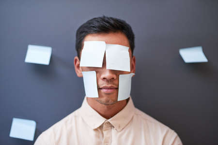 An Asian businessman stands with sticky notes on his face looking tired at workの写真素材