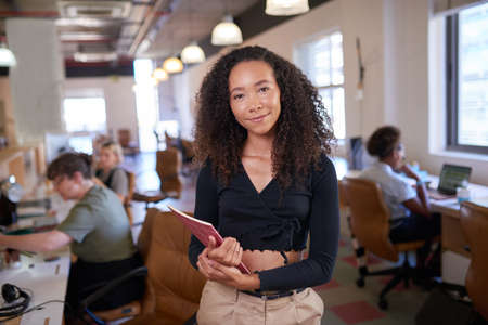 A multi-ethnic woman stands in office with team members in backgroundの写真素材
