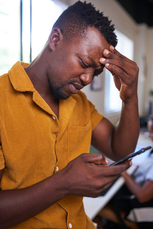 A Black man rubs his forehead looking confused while on his mobile deviceの写真素材
