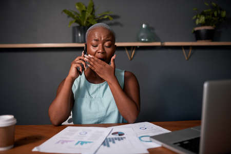 A Black businesswoman yawns at her office desk late at night reading reportsの写真素材