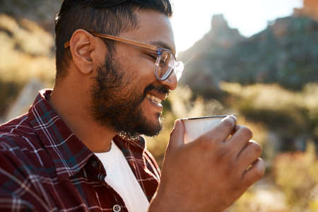 A South Asian man smiles as he sips coffee on camping trip in the mountainsの写真素材
