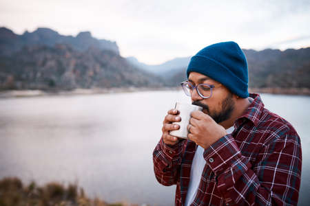 A South Asian man sips coffee from a camping mug with mountains and lake viewの写真素材