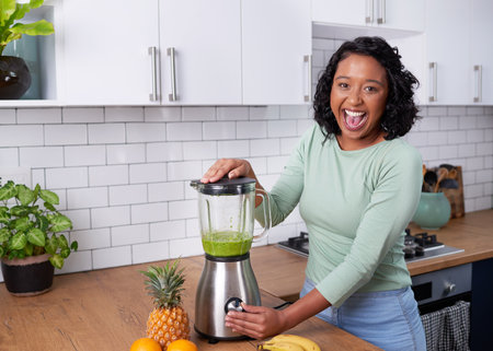 An excited young woman blends green smoothie in the kitchen at homeの写真素材