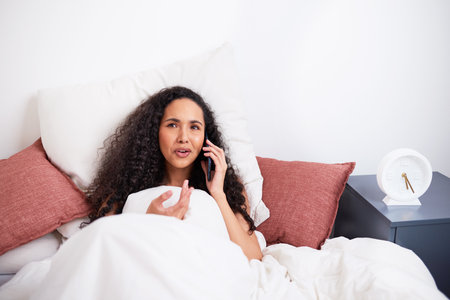 A young multi-ethnic woman gestures mid-conversation during call in bedの写真素材