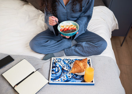 Cropped overhead shot of young woman eating breakfast in bed with journal