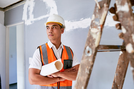 A serious construction worker stands with plans under his arm at home renovationの写真素材