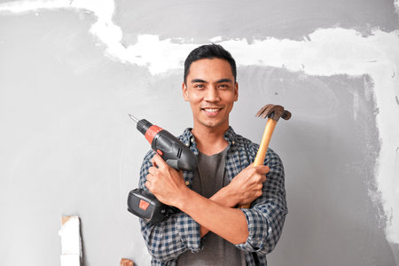 A young Asian man smiles while holding drill and hammer for home DIY improvementの写真素材