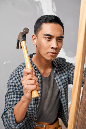 A serious Asian man uses hammer to hit wood while doing home renovation DIY