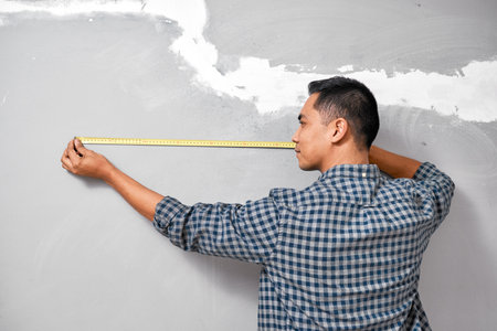 An Asian man measures against the wall using measuring tape doing home DIYの写真素材