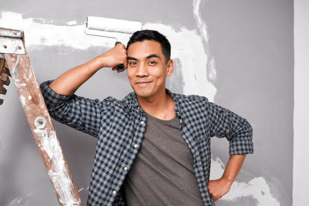 An attractive Asian man smiles and poses with paint roller against wallの写真素材