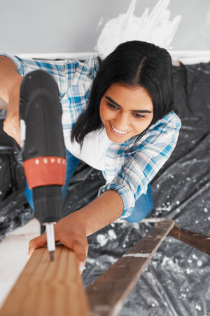 Overhead shot of young Indian woman drilling into wood during home renovationの写真素材