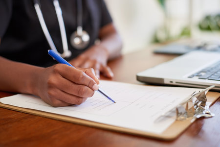 Close up shot of Black doctor filling in patient symptoms on clipboard at desk