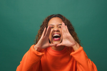 Young biracial woman raises hands, yells excitedly, green background studioの写真素材
