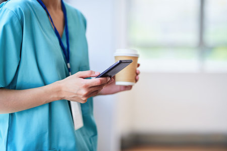 Medical Professional in Scrubs Using Smartphone While Holding Coffee in Hospitalの写真素材