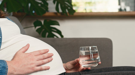 Pregnant Woman Relaxing on Couch Holding Water Glass for Healthy Hydration and Comfort at Homeの写真素材