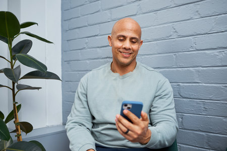 Man in Casual Clothing Smiling While Using Smartphone, Seated in a Cozy Indoor Setting