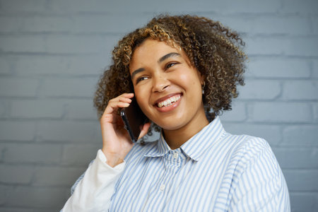Woman Talking on Phone Smiling in Casual Outfit Against Brick Wall Backgroundの写真素材