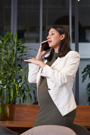 Pregnant Businesswoman Taking a Phone Call in a Modern Office Setting, Wearing a White Blazer and Green Dressの写真素材