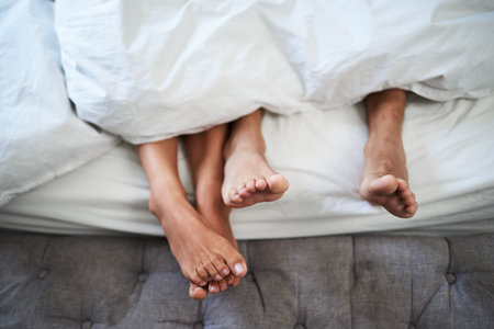 Close-Up of a Couples Relaxed Feet Peeking Out from White Bedding on a Cozy Bedの写真素材