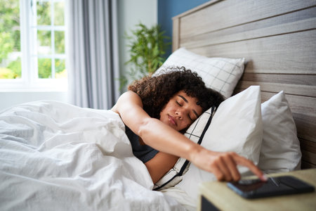 Young Woman Lying in Bed Reaching for Smartphone on Nightstand in Morningの写真素材