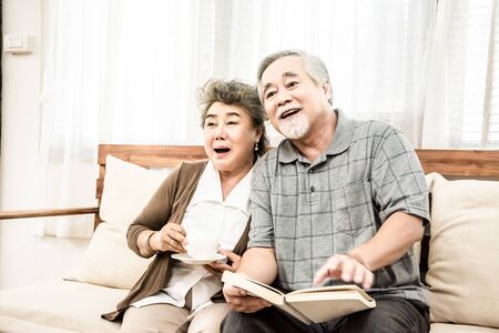 Asian elderly couple sitting together in the living room reading a book and Drinkingの写真素材