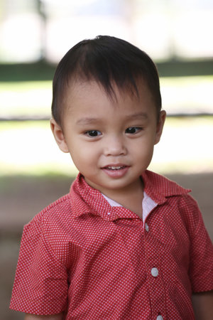 An Asian boy in a red shirt is smiling happily on blurred scenery background.の写真素材