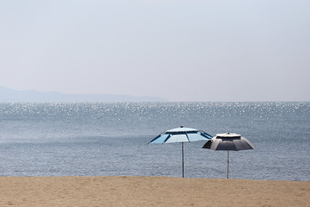 Two beach umbrellas spread out on the beach on a hot sunny day, Beach and the sea with the sunlight reflecting on the sparkling sea water.の写真素材