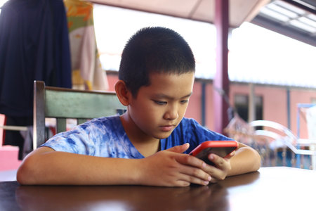 A young boy is focused on his smartphone while sitting at a table in a cozy indoor setting, representing the modern youth's engagement with technology.の写真素材