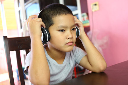 A young boy sits at a table, wearing headphones, deeply engaged in listening to music. His focused...