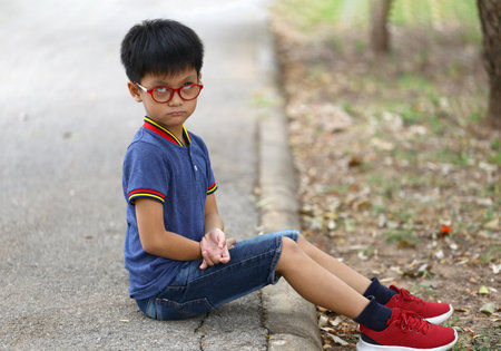 A young boy with glasses sits thoughtfully on a pathway, showcasing his casual summer style. The serene outdoor setting enhances his contemplative mood.の写真素材