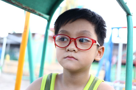 A thoughtful young child wearing bright red glasses sits at a playground. The image captures a moment of introspection, showcasing childhood innocence and curiosity in a vibrant setting.の写真素材