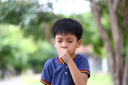 A young boy shows a concerned expression while coughing outdoors. The natural backdrop highlights...