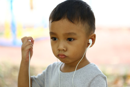 A young boy wearing earphones enjoys music in a sunny outdoor park. His thoughtful expression captures the essence of childhood curiosity and joy.の写真素材