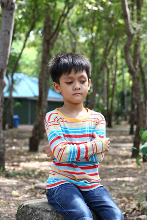A thoughtful young boy sits outdoors in a peaceful forest, surrounded by trees. His colorful...