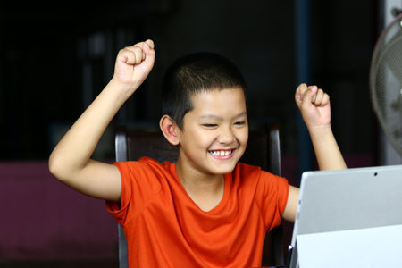 A joyful child celebrates a victory while using a tablet. His excited expression showcases pure happiness in a casual indoor setting. Perfect for themes of technology and childhood joy.の写真素材