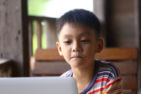 A young boy sits indoors, focused on his laptop. This image captures his concentration and curiosity, reflecting the modern learning environment and technology's role in childhood development.の写真素材