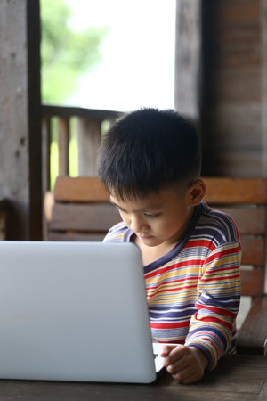 A young boy sits outdoors on a wooden bench, focused on his laptop. He wears a colorful striped shirt, surrounded by natural light and a cozy atmosphere.の写真素材