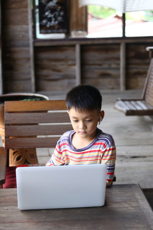 A young boy sits at a wooden table, intently using a laptop. The cozy indoor setting, including wooden beams and furniture, enhances the atmosphere of focused learning and digital engagement.の写真素材