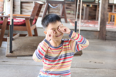 A joyful child sits on a swing, playfully covering his eyes with a big smile. This charming indoor scene captures the essence of carefree childhood moments filled with happiness.の写真素材