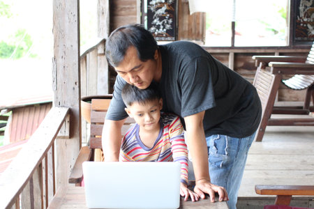 A father and his son engage with a laptop at home, showcasing a moment of bonding and learning in a cozy indoor setting filled with warmth.の写真素材