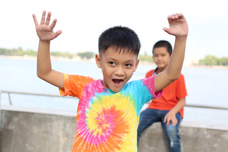 A joyful child wearing a colorful tie-dye shirt celebrates by the waterfront, radiating happiness and excitement in a vibrant outdoor setting.の写真素材