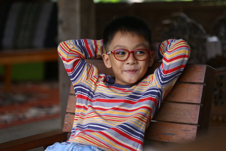 A relaxed young boy sitting comfortably on a wooden chair, wearing a colorful striped sweater and...