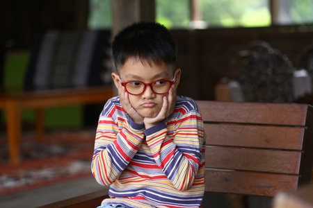 A thoughtful boy with glasses sits indoors, resting his chin on his hands. The cozy atmosphere creates a perfect backdrop for reflection and emotion.の写真素材