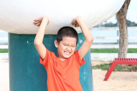 A joyful boy in an orange shirt plays energetically at a park, lifting a large sculpture with a big smile, capturing the essence of childhood happiness and fun.の写真素材