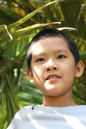 A joyful young boy smiles confidently while standing under lush green leaves, radiating innocence and happiness in the warm sunlight. Perfect for nature-focused themes.の写真素材